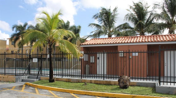 Outdoor swimming pool area surrounded by a fence, with a building and palm trees in the background.