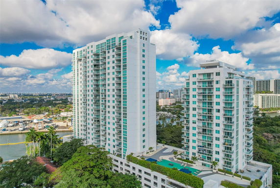 Aerial view of two modern high-rise residential buildings with glass balconies, surrounded by a cityscape and a river nearby.
