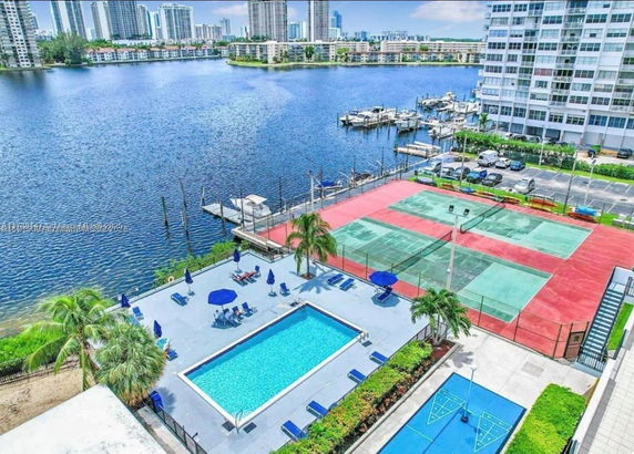 View of tennis courts and a swimming pool next to a body of water with residential buildings in the background.