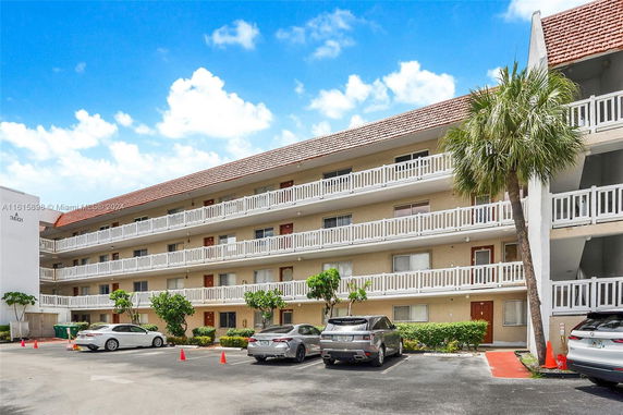 View of a multi-story building with a parking lot in front, featuring several cars parked and a palm tree nearby.
