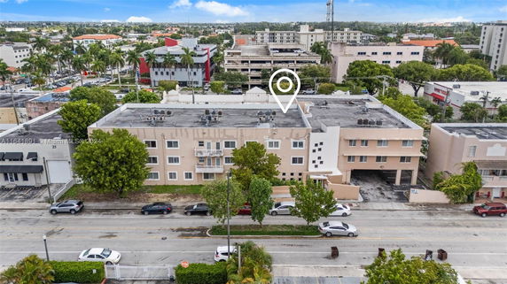 Aerial view of a multi-story building with a flat roof and visible AC units, surrounded by other buildings and a tree-lined street.