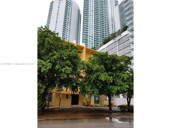 Front view of a residential building with a yellow exterior, surrounded by taller modern apartment buildings.