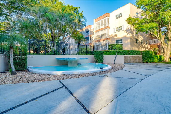 View of a multi-story building with balconies surrounded by trees and a water feature.