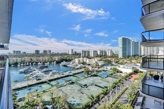 Panoramic view from a high-rise balcony showing a marina with boats, multiple tennis courts, and tall buildings in the background.