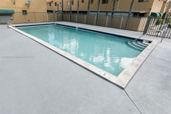 Outdoor swimming pool with surrounding fence and apartment buildings in the background.