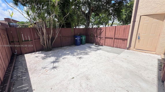 Outdoor area with wooden fencing, a concrete floor, and some green trash bins.