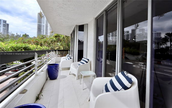 Outdoor balcony with white chairs and blue-striped cushions, overlooking surrounding buildings and greenery.