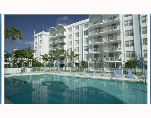 Outdoor swimming pool area in front of a multi-story white building with balconies.