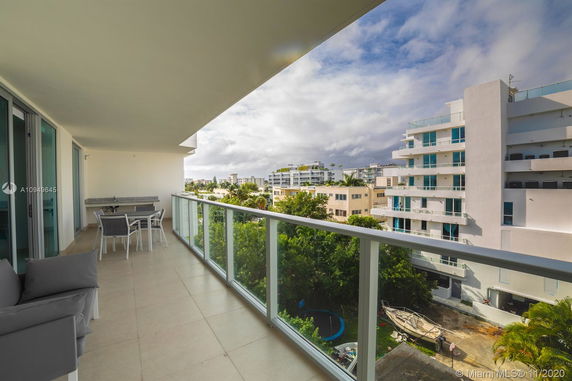 Balcony view from a high-rise building with glass railing and outdoor furniture, overlooking nearby modern apartment buildings.