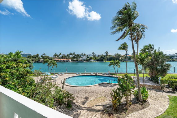 View of an outdoor swimming pool area with palm trees and water in the background.