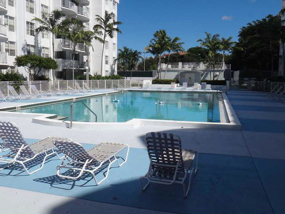 Outdoor swimming pool area with surrounding lounge chairs and a multi-story building in the background.