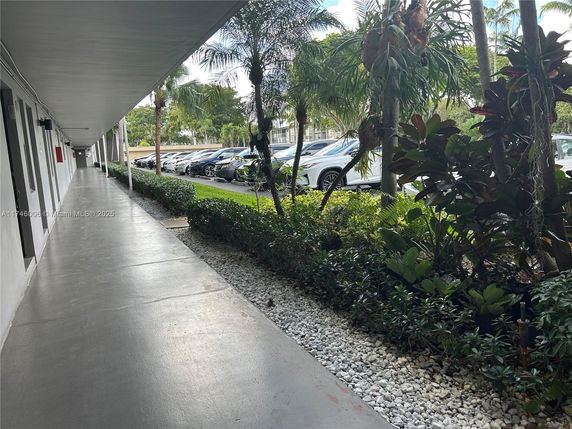 View of a covered walkway next to an outdoor parking area with cars and surrounding greenery.