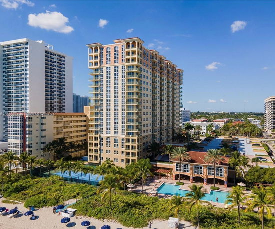 View of a multi-story residential building with balconies, located near a beach. Surrounding areas include other tall buildings and a swimming pool.