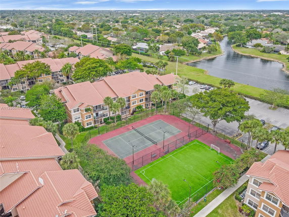 Aerial view of apartment buildings with tennis court and green field, surrounded by trees and a nearby lake.