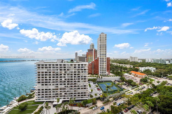 View of a waterfront multi-story residential building with nearby tennis courts and high-rise structures in the background.