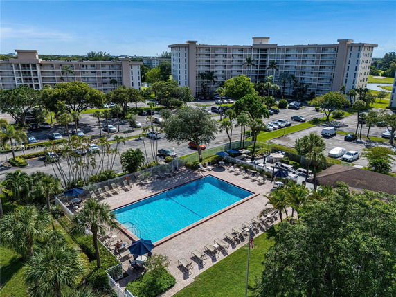 Outdoor swimming pool area with lounge chairs, surrounded by trees and parking, in front of multi-story residential buildings.