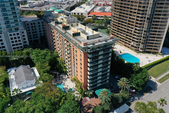 Aerial view of a tall apartment building with a flat rooftop containing various structures and equipment. Surrounding areas include greenery and nearby buildings.