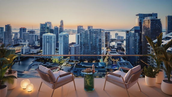 Panoramic view of a cityscape with skyscrapers and a water body at sunset, seen from a balcony with two chairs and plants.