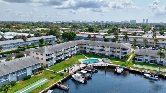 Aerial view of a residential complex near a waterfront with boats docked nearby, featuring multiple buildings with balconies and a swimming pool.