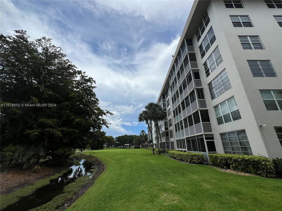 Angled view of a multi-story residential building with screened balconies and numerous windows. The surroundings include a grassy area, trees, and a small waterway.