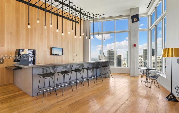 Modern indoor space with a long bar counter and tall stools, featuring large windows with city views.