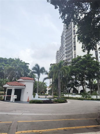 Front view of a tall residential building with balconies, surrounded by trees and a guardhouse.