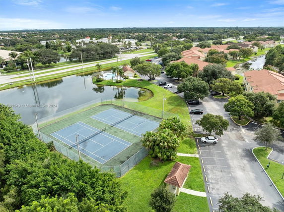 Aerial view of tennis courts adjacent to a parking area and a small pond in a residential or community setting.