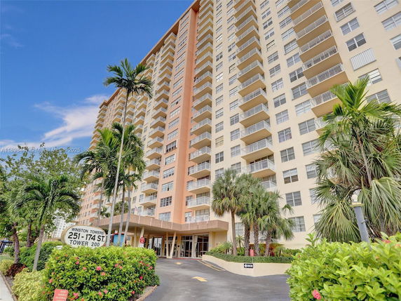 Front view of a tall residential building with balconies and palm trees in front.