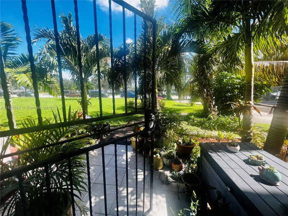 View from a porch or balcony with metal railing, overlooking a garden area with palm trees and potted plants.