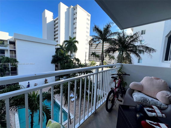 View from a balcony overlooking a swimming pool and nearby apartment buildings with palm trees.