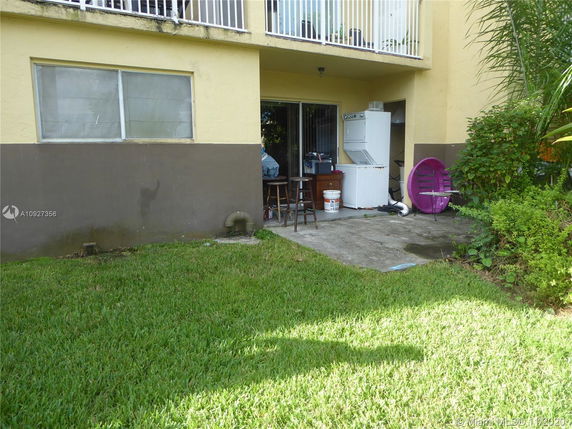 View of a building exterior with a grassy area, showing a ground-floor patio with a washing machine and some outdoor furniture.