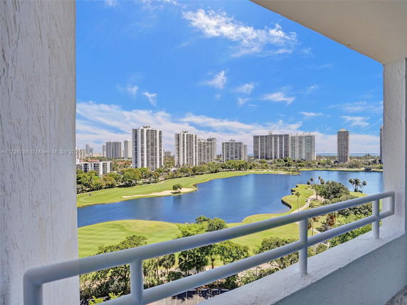 Panoramic view from a building showcasing a lake, a golf course, and tall city buildings under a clear blue sky.