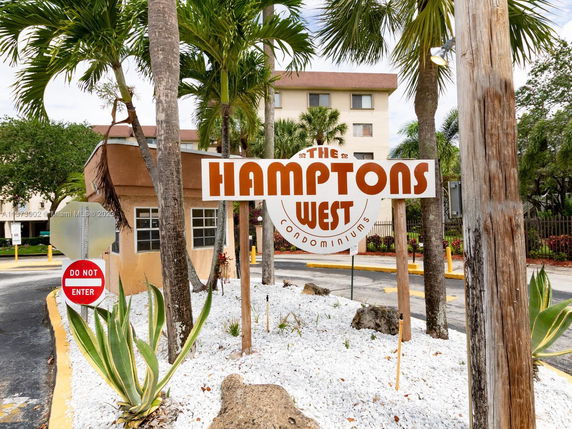 Front view of a condominium building with a 'Hamptons West' sign and palm trees.