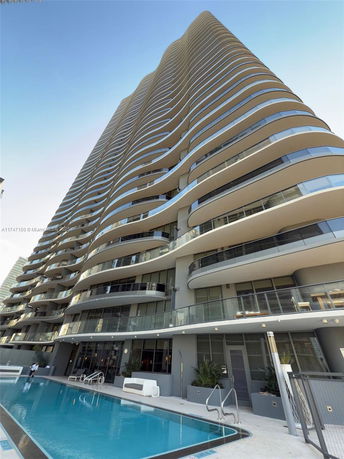 Front view of a tall, modern residential building with curved balconies and a swimming pool at the base.
