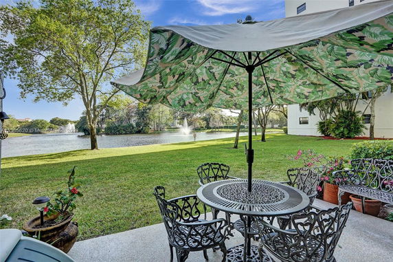 Patio area with metal furniture and parasol, overlooking a pond with a fountain and trees.
