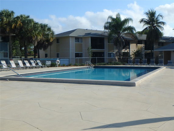 Outdoor swimming pool area with lounge chairs and a multi-story building in the background.