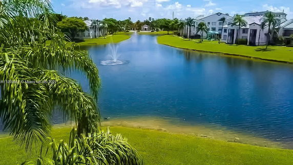 View of a pond with a fountain, surrounded by greenery and houses.