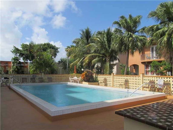 View of a swimming pool area with palm trees and a building nearby.