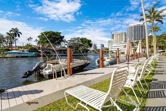 View of a dock with boats anchored by a waterway, surrounded by buildings and palm trees.