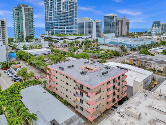 Aerial view of a mid-rise apartment building with pink and beige exterior in a cityscape with nearby skyscrapers and ocean in the background.