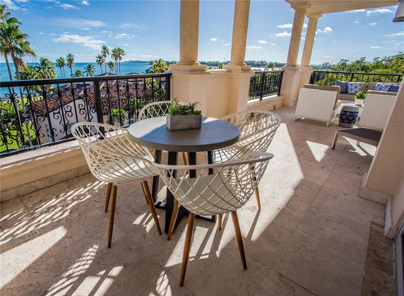 Balcony with white lattice chairs and round table overlooking ocean and palm trees.
