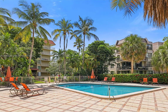 Outdoor swimming pool area with palm trees and apartment buildings in the background.