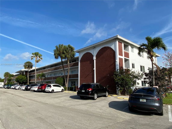 Front view of a three-story building with multiple balconies and a parking area in front.