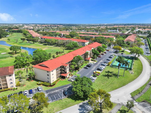 Aerial view of residential complex with parking area and tennis courts, surrounded by green spaces.