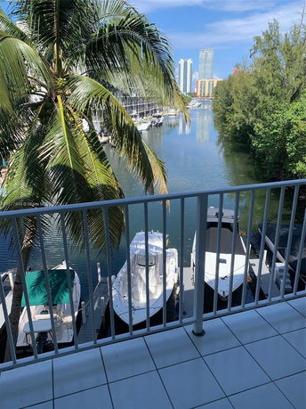 View of boats docked on a waterway with tall buildings and trees in the background, seen from a balcony.