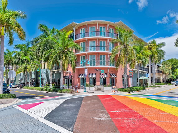 Front view of a multi-story building with balconies and storefronts, located at a street corner with palm trees nearby.