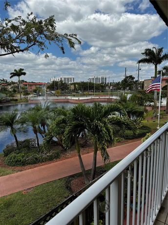 View from a building balcony overlooking a landscaped area with palm trees, a pond, and distant buildings under a cloudy sky.