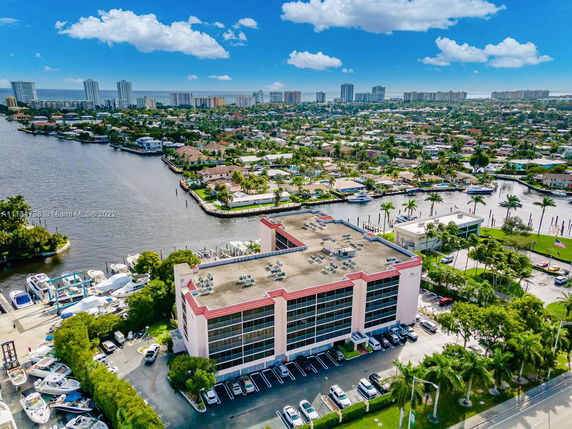 Aerial view of a multi-story building near a waterway with residential and commercial areas in the background.