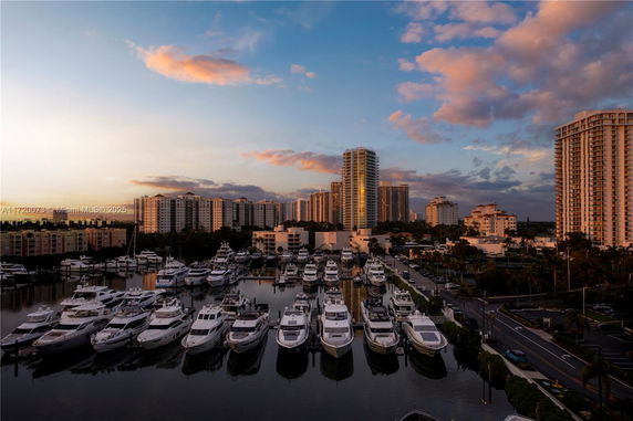 Panoramic view of a marina with numerous yachts docked, surrounded by high-rise buildings under a colorful sky.