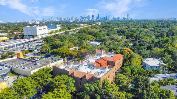 Aerial view of a brown apartment building with a red-tiled roof surrounded by trees, with a city skyline in the background.